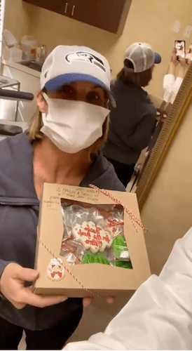 Woman wearing face mask, baseball cap, holding a box of sugar cookies.